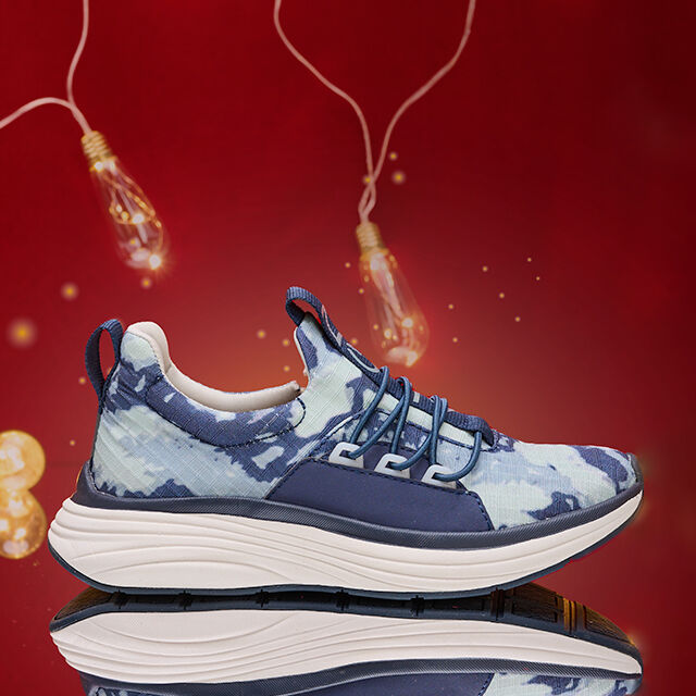 Zapatilla sin cordones Theora azul para mujer en Tie Dye, sobre un fondo rojo con luces encendidas en el fondo, tomada en un espejo